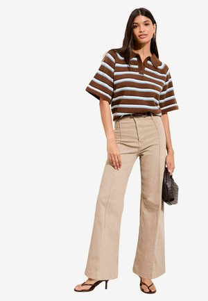 Nő barna és világoskék csíkos rövid ujjú pulóvert, bézs magas derekú, széles szárú nadrágot, fekete sarkú szandált visel, és egy texturált sötét kézitáskát tart a kezében.