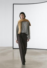 Haut gris à manches longues avec une poche poitrine, associé à un pantalon droit marron foncé. Une écharpe en maille marron clair est drapée sur les épaules.