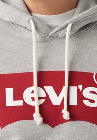 Szara bluza z kapturem z czerwonym logo Levi's. Posiada białe sznurki do ściągania oraz miękką teksturę. Projekt zawiera kontrastującą, zaokrągloną dolną krawędź.