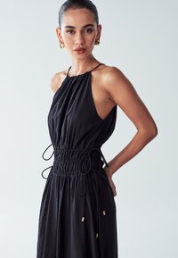Black sleeveless dress with gathered neckline, adjustable drawstring waist, and gold accent details at the waist. Smooth fabric texture.