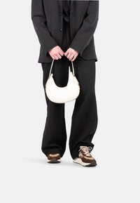 Borsa curva bianca tenuta in mano, con un accento di hardware dorato. Indossata con un blazer nero oversize e pantaloni neri larghi. Sneakers marroni.