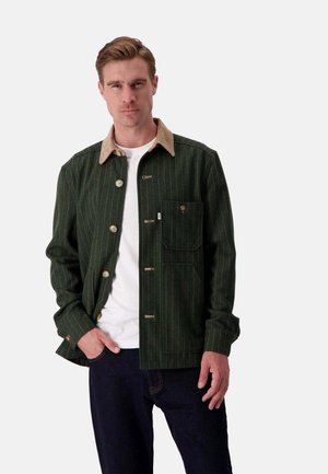 Dunkelgrüne gestreifte Jacke mit einem hellbraunen Cordkragen, vorderen Knöpfen und einer Brusttasche. Getragen über einem weißen Hemd und dunklen Jeans.