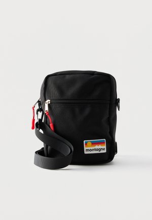 Petit sac bandoulière noir compact avec poche zippée à l'avant, tirettes rouges, sangle réglable et écusson coloré avec le logo "montagne".