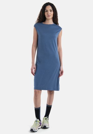 Blauwe, knielange jurk met kortemouwen, gemaakt van zachte stof. Gekleed met grijze sneakers met gele accenten en zwarte sokken.
