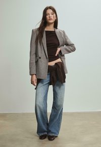 Blazer à carreaux, haut ajusté marron, pull marron noué, jeans larges bleu clair, et chaussures marron foncé. Textures lisses et couleurs contrastées.