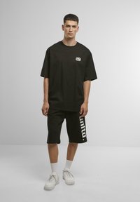 Black oversized t-shirt with a small logo on the chest, paired with black shorts featuring a white "unita." text, completed with white sneakers.