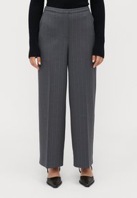 Vrouw in hoog aangesloten grijze gestreepte wijde broek, zwarte lange mouwen top en zwarte puntige schoenen, staand tegen een effen achtergrond.