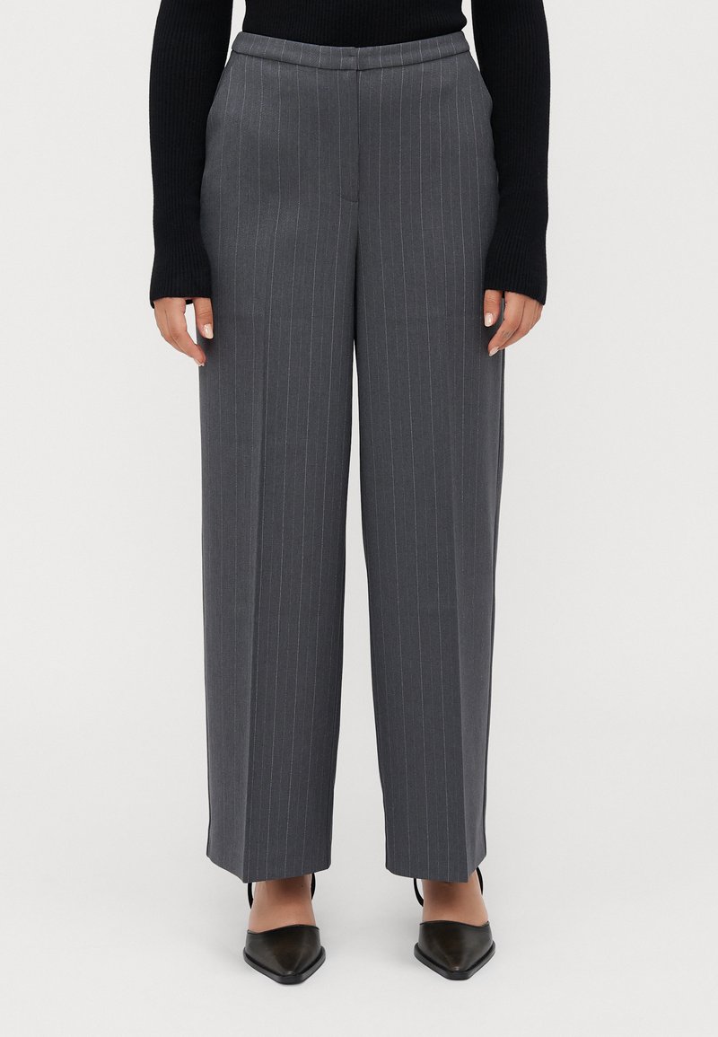 Vrouw in hoog aangesloten grijze gestreepte wijde broek, zwarte lange mouwen top en zwarte puntige schoenen, staand tegen een effen achtergrond.