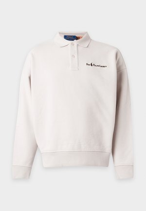 Mann trägt weißes langärmeliges Polohemd, beige Cargohosen und beige Loafers, steht vor einem schlichten hellen Hintergrund.