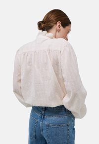 Blouse rose clair en tissu texturé, arborant des motifs subtils, un col volanté et des manches bouffantes, accompagnée d'un jean en denim bleu.