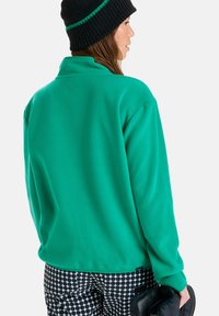 Pullover de fleece azul-água com colarinho dobrável, corte descontraído e mangas longas; utilizado com calças pretas estampadas e um gorro preto listrado.