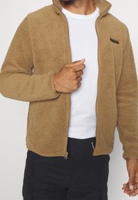 Tan fleecejacka med hög krage och full dragkedja. Har en texturerad yta, sidofickor och logotyp på bröstet.