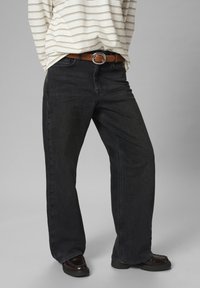 Osoba u širokim crnim trapericama, smešnim kaišem s srebrnom kopčom, prugastoj košulji s dugim rukavima i tamnosmešnim cipelama stoji uz sivu pozadinu.