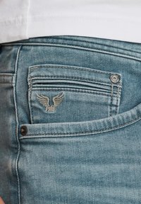 Jeans de mezclilla azul claro con un logo alado bordado en el bolsillo trasero y un bolsillo horizontal adicional encima de este.