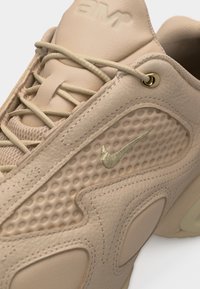 Šviesiai rudos spalvos Nike sportbačiai su tekstūruotu šoniniu paneliu, odiniais sluoksniais, šviesiai rudos spalvos raišteliais, auksiniu skylutės žiedu ir įspaustu Nike logotipu liežuvėlyje.