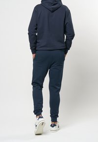 Tmavě modrá mikina s kapucí a odpovídající tepláky. Měkká látka, volný střih, žebrované manžety a klokaní kapsa vpředu.