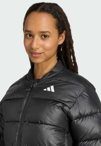 Mladá žena s vrkôčikmi sa usmieva, oblečená v čiernej prešívanej bunde s bielym logom. Pozadie je svetlo šedé.