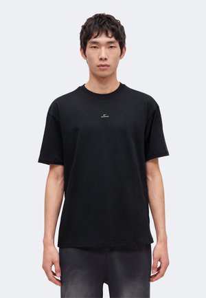 Junger Mann mit kurzen dunklen Haaren, der ein schlichtes schwarzes Nike-T-Shirt und schwarze Hosen trägt und vor einem hellgrauen Hintergrund steht.