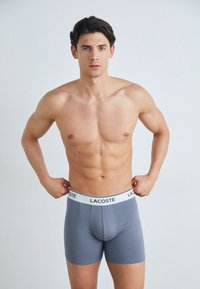 Grijze katoenen boxershorts met een witte Lacoste tailleband, met een strakke pasvorm en een gladde textuur. Model staat rechtop en past de tailleband aan.