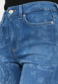 Μπλε τζιν παντελόνι με ελαφρύ λουλουδάτο μοτίβο, με κλασικό σχεδιασμό πέντε τσεπών, ορειχάλκινο κουμπί και διακριτική λεπτομέρεια με ξεθώριασμα.