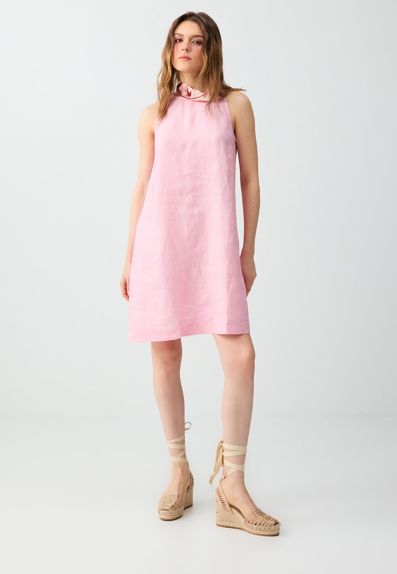Jeune femme portant une robe rose sans manches avec un col foulard et des espadrilles compensées beiges avec des liens à la cheville, debout devant un fond uni et clair.