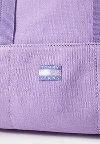 Lila Leinen-Einkaufstasche mit einem blauen rechteckigen Etikett, auf dem "TOMMY JEANS" steht. Die Tasche verfügt über robuste Nähte und ein strukturiertes Material.
