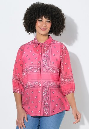 Kvinde med krøllet hår iført en lyserød paisley-mønstret skjorte med 3/4-ærmer og blå jeans, smilende mod en hvid baggrund.