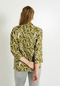 Veste vert olive avec un motif de feuilles, manches trois-quarts et coupe décontractée. Le matériau semble léger avec une finition texturée.