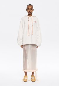 Sudadera blanca de gran tamaño con cordones trenzados, combinada con una falda beige transparente con un dobladillo con volantes y zapatos de cordones color carne.