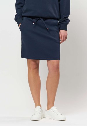 Persoon die een marineblauwe knielange rok met zakken en witte sneakers draagt, staand tegen een effen lichte achtergrond.