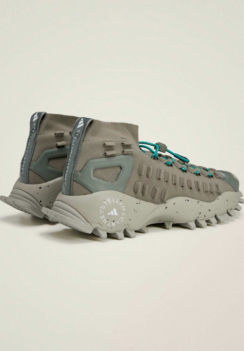 Grijze sportschoenen met een gebreide constructie, asymmetrisch ontwerp, teal accenten en een robuuste, gestructureerde zool met opvallende traction studs.