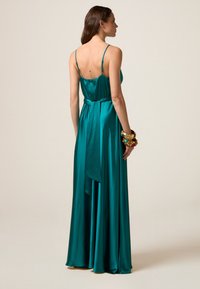 Abito lungo in satin color teal con spalline sottili, corpino aderente e gonna fluida. Presenta un punto vita legato e un dettaglio drappeggiato sulla schiena. Bangles dorati al polso.