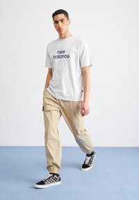 Weißes Kurzarm-T-Shirt mit "new balance"-Logo, kombiniert mit beigen Cargo-Hosen mit seitlichen Taschen und schwarz-weißen karierten Sneakers.
