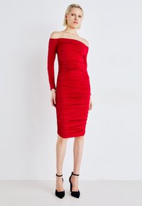 Rochie roșie off-shoulder din material elastic, cu detalii pliate, siluetă fitted și tiv până la genunchi. Asortată cu tocuri negre.