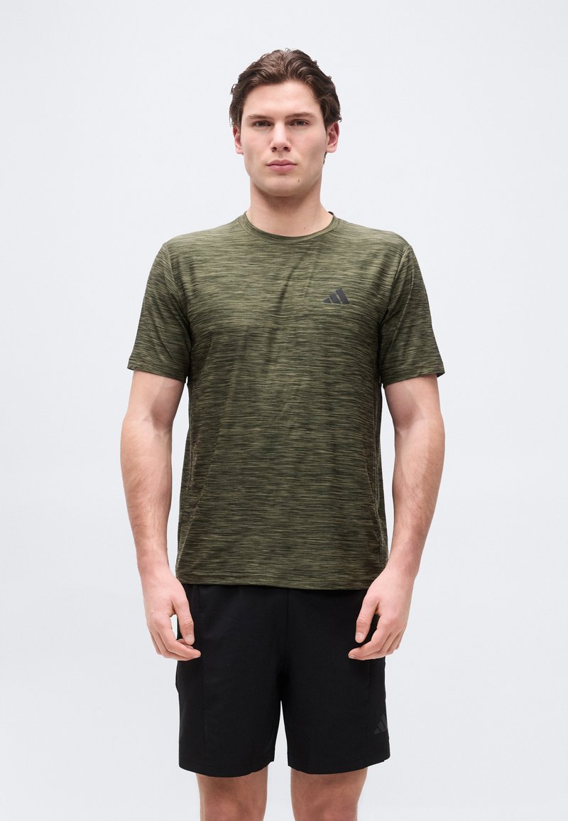 Άνδρας που φοράει αθλητική μπλούζα Adidas σε ελιά πράσινο και μαύρο σορτς, στέκεται με τα χέρια χαλαρά σε ένα απλό φωτεινό φόντο.