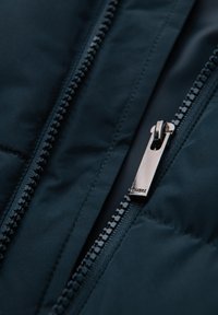 Navyblauwe gewatteerde jas close-up, met een soepele metalen rits met merkimprint en textuurdetail langs de ritsranden.