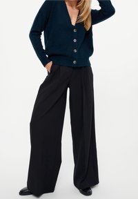 Mörkgrön stickad cardigan med V-ringning och silverknappar, kombinerad med svart midjehög vida byxor. Slät textur, avslappnad passform.