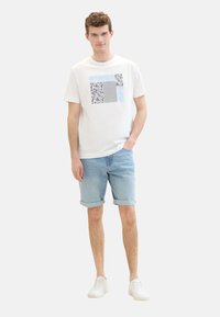Biela bavlnená tričko s grafickým dizajnom v modrej a čiernej farbe, skombinované s svetlými denimovými šortkami. Biely tenisky. Jednoduchý, neformálny outfit.