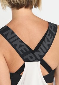 Dos d'une femme montrant les bretelles croisées noires d'un soutien-gorge de sport Nike avec le nom de la marque visible, sous un débardeur athlétique blanc.