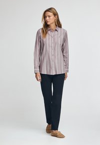 Camicia a maniche lunghe, con bottoni, color marrone chiaro con righe verticali bianche. Abbinata a pantaloni scuri e mocassini beige. Tessuto dalla finitura liscia.