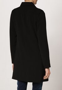 Černý kabát s límcem, dlouhými rukávy a rovného střihu. Hladká textura, délka ke kolenům, vybavený bočními kapsami.