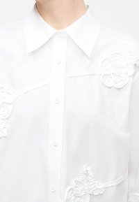 Chemise blanche à boutons avec col pointu, présentant une broderie florale texturée sur les zones de la poitrine et des épaules.