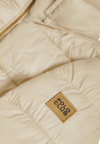 Veste puffer beige avec texture matelassée, dotée d'une étiquette de marque avec accents dorés. Fermeture éclair et tissu lisse et brillant.