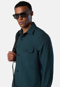 Camisa verde de botones con un bolsillo en el pecho, mangas largas y un parche de logo negro en el hombro izquierdo. Llevada con gafas de sol oscuras.