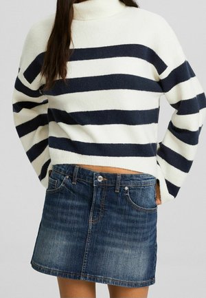 Kvinde iført en hvid og navyblå langærmet trøje med striber samt en blå denim mininederdel med forlommer og knaplukning.