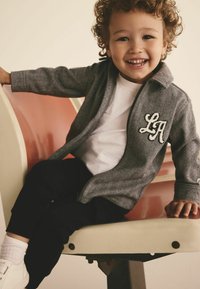 Young child with curly hair smiling, wearing a gray jacket with "LA" letters, white shirt, black pants, and white shoes, sitting on a bench.