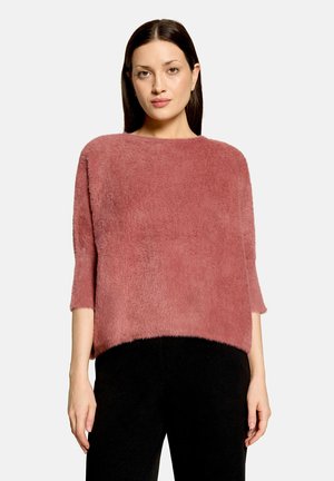 Femme aux cheveux raides et foncés portant un pull ample et duveteux de couleur rose et un pantalon noir, debout devant un fond uni.