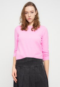 Ροζ εφαρμοστό πουλόβερ σε μαλακό υλικό με μακριά μανίκια, λαιμόκοψη στρόγγυλη και ελαφρύ rib ύφανση στις μανσέτες, συνδυασμένο με σκούρα φούστα με πιέτες.