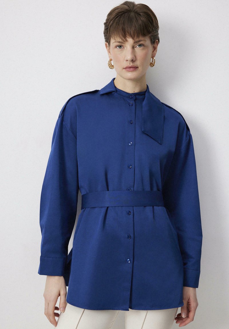 Camicia blu a maniche lunghe con colletto, dettaglio cravatta e vita cintrata. Tessuto liscio con una leggera lucentezza, con bottoni frontali.