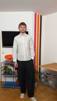 Νεαρός άνδρας με λευκό πουκάμισο με ρίγες και μαύρο παντελόνι στέκεται σε δωμάτιο με πολύχρωμο τοίχο με ρίγες, πορτοκαλί φωτιστικό, τηλεόραση, φυτά και βαλίτσα με αυτοκόλλητα.